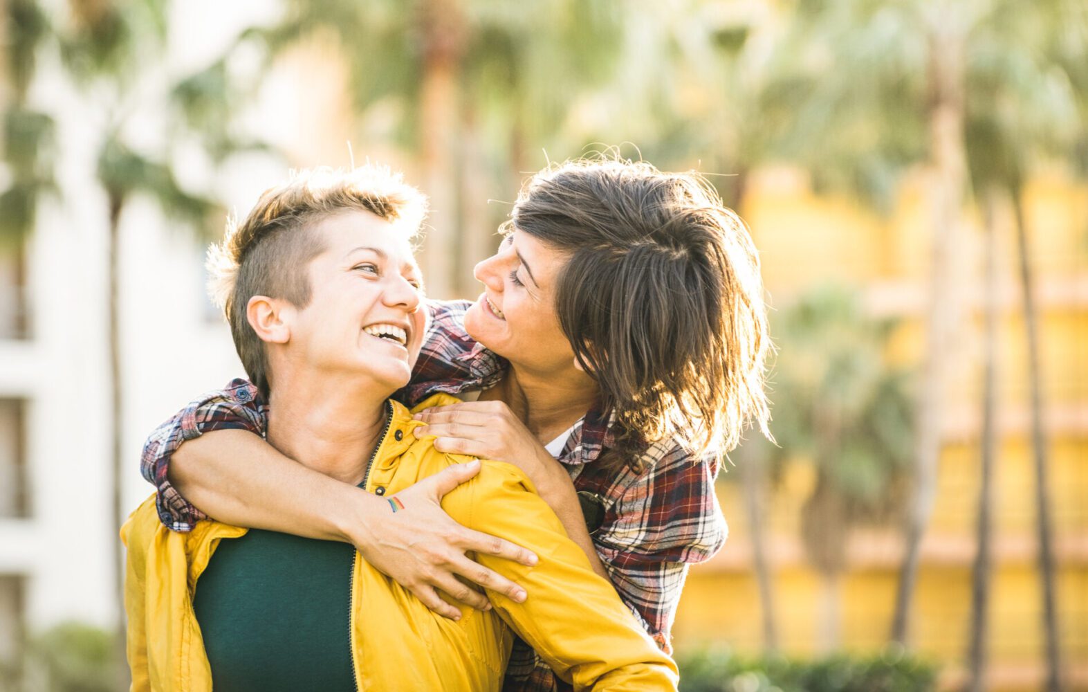 Happy playful girlfriends in love sharing time together at travel trip on piggyback hug - Women friendship concept with girls couple having fun on fashion clothes outdoors - Bright warm sunset filter